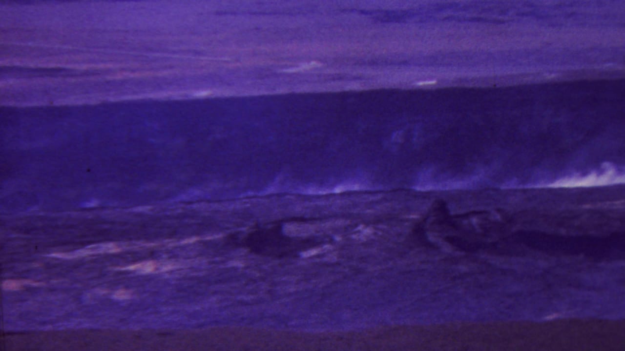 1962-attributed Hawaii home-movie frame with dim purple terrain, a dark central form, and faint pale plume or surf marks under low-light exposure.