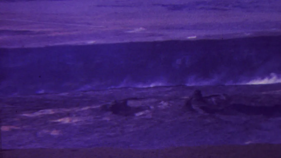 1962-attributed Hawaii home-movie frame with dim purple terrain, a dark central form, and faint pale plume or surf marks under low-light exposure.