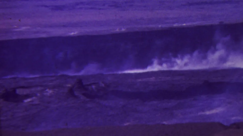 1962-attributed Hawaii frame with bright irregular steam or surf-like plumes crossing a dark purple volcanic or coastal landscape, rendered in grainy 8mm color.