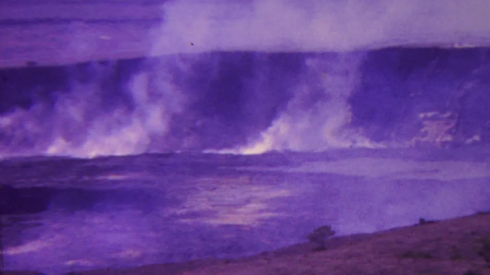 1962 Hawaii 8mm frame with smoke or steam billowing near the waterline, reflective surf below, and a barren sloped volcanic shoreline in the foreground.