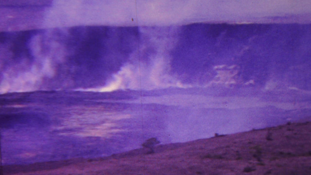1962 Hawaii 8mm frame showing a smoky volcanic crater beyond rough dark terrain, with strong purple color shift across the landscape.