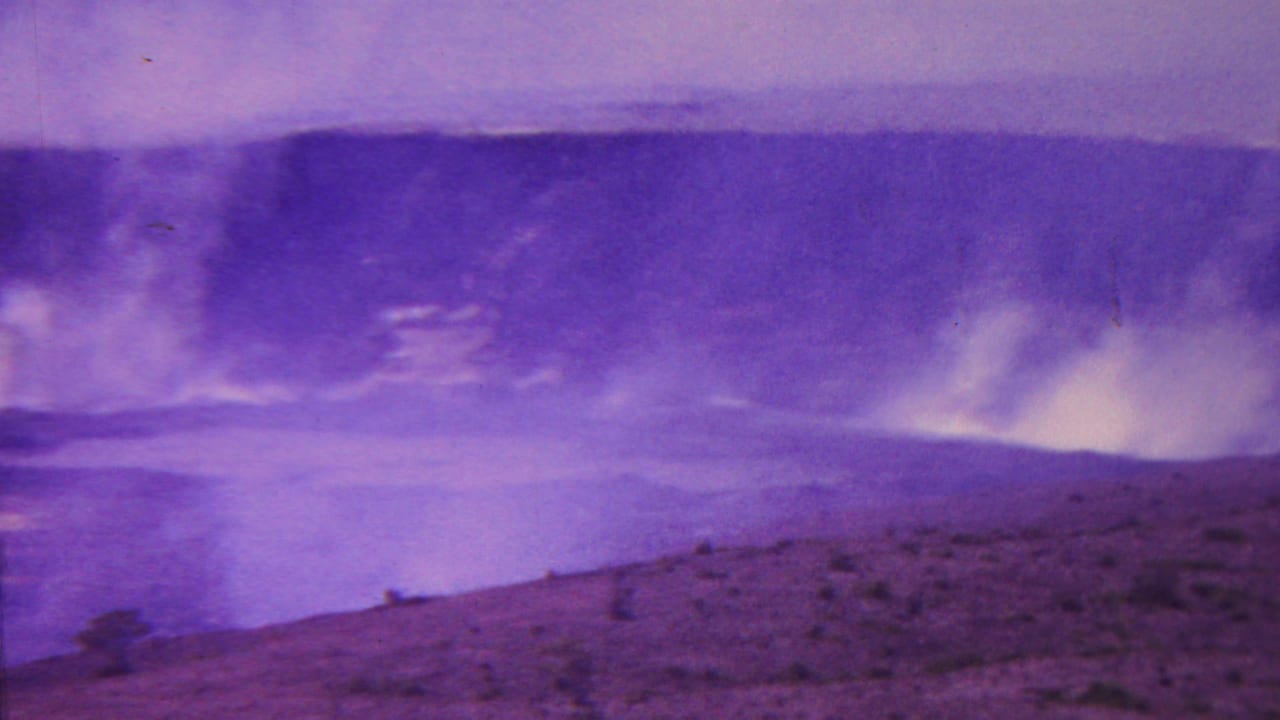 1962 Hawaii 8mm frame with a broad view of crater smoke, pale glowing areas near the base, and rocky volcanic ground in the foreground.