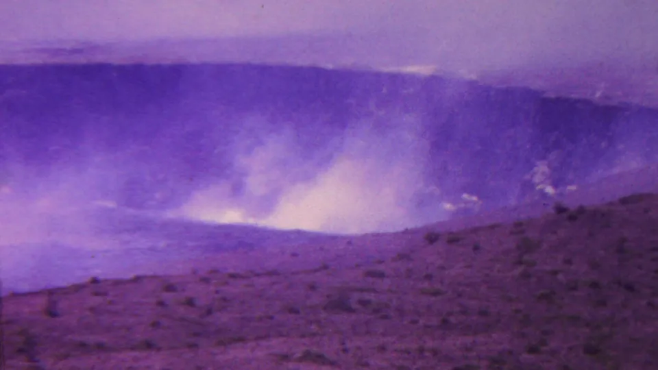 1962 Hawaii 8mm frame showing bright smoky haze inside a volcanic crater behind sloped, rough volcanic ground, rendered with heavy purple color shift.