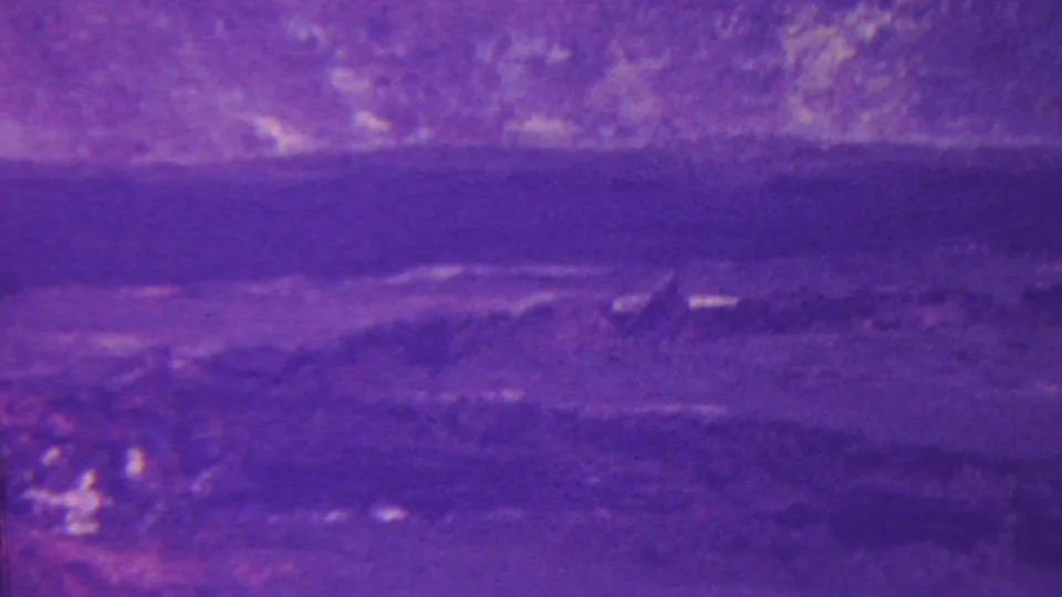 1962 Hawaii home-movie frame showing dark foreground terrain, a hazy canyon floor, and lighter textured slopes or cliffs under a strong purple film shift.