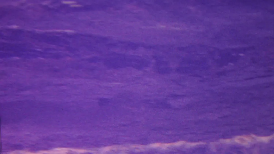 1962 Hawaii home-movie frame with a faded purple view across a canyon or valley floor toward sloping terrain and pale rock or vegetation detail.