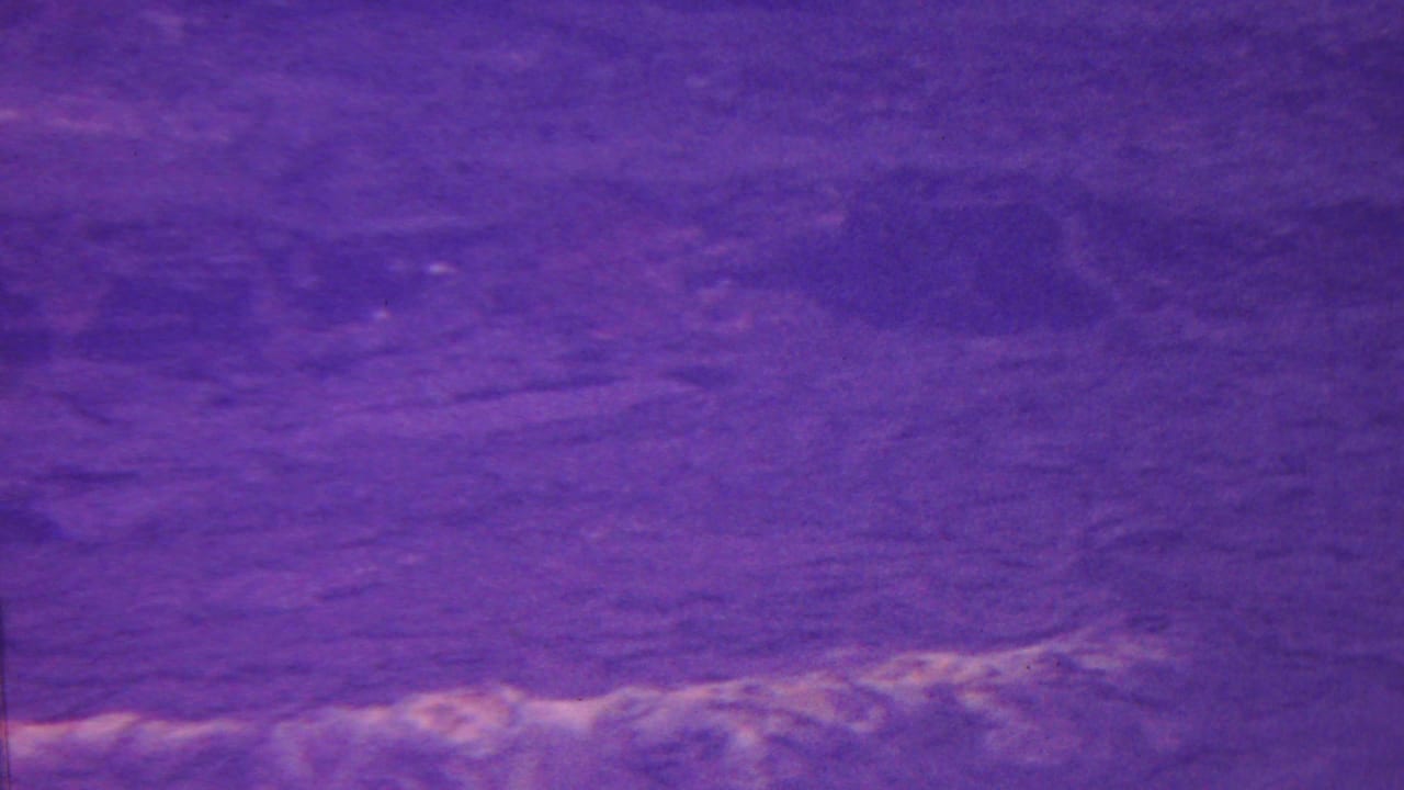 Purple-tinted 1962 Hawaii 8mm frame showing a close view of rippled water or wet ground with soft pale reflections and dark surface patches.
