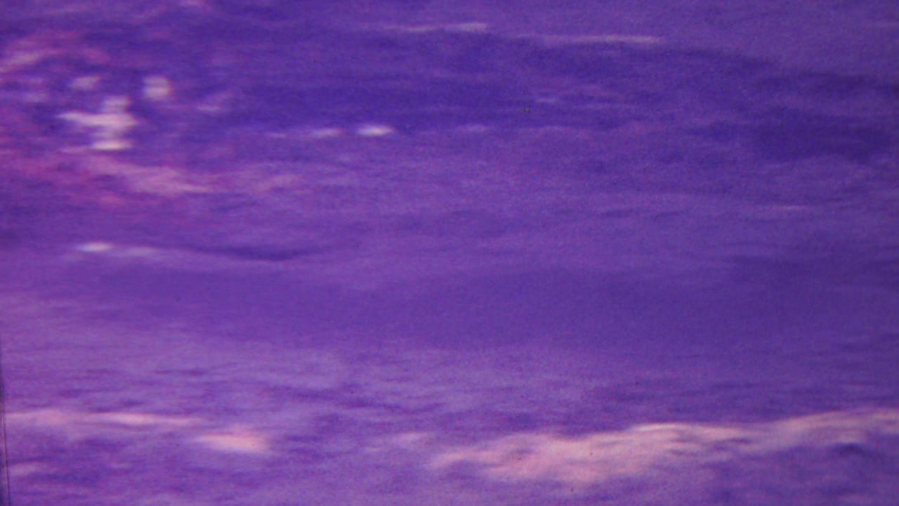 1962 Hawaii home-movie frame with a purple-blue cast, showing ripples, reflected streaks, and indistinct wet ground or water texture.