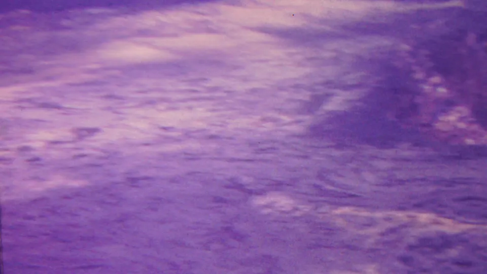 Purple-tinted 1962 Hawaii 8mm frame showing uneven wet ground or shallow water with pale reflections, darker speckling, and analog softness.