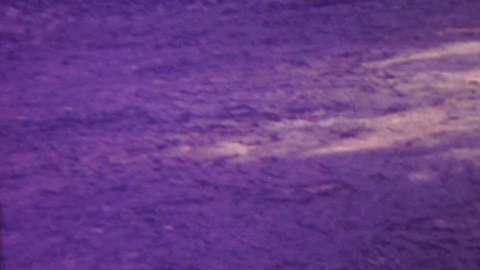 1962 Hawaii home-movie frame with purple toning, showing rippled ground or water texture and a curved pale streak across the right side.