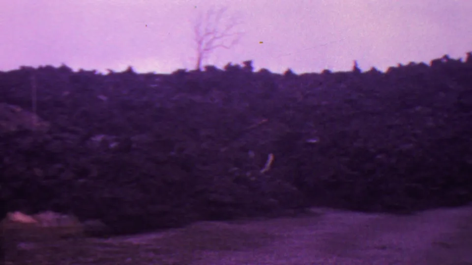 1962 Hawaii 8mm frame showing a dark burned slope or brush line beneath a hazy purple-tinted sky, with pale ground in front and a bare tree shape near the horizon.