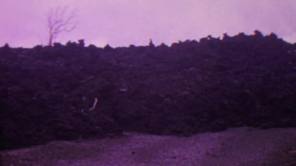 1962 Hawaii 8mm frame with a static view of dark burned earth or brush, pale bare ground at the lower edge, and a soft haze across the purple-shifted sky.