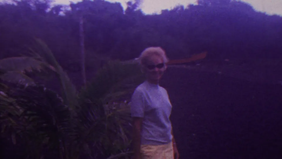 An adult woman with short blonde hair and sunglasses stands near tropical plants and a dark pond or water area in the 1962 Hawaii home movie.