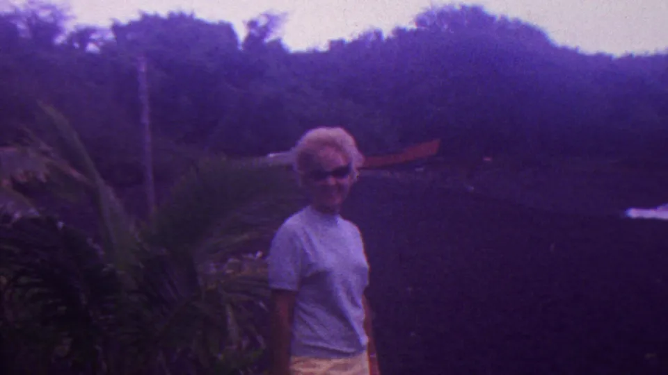 In supplied 1962 Hawaii 8mm footage, a woman with short blonde hair and sunglasses stands near palm-like plants and dense greenery.