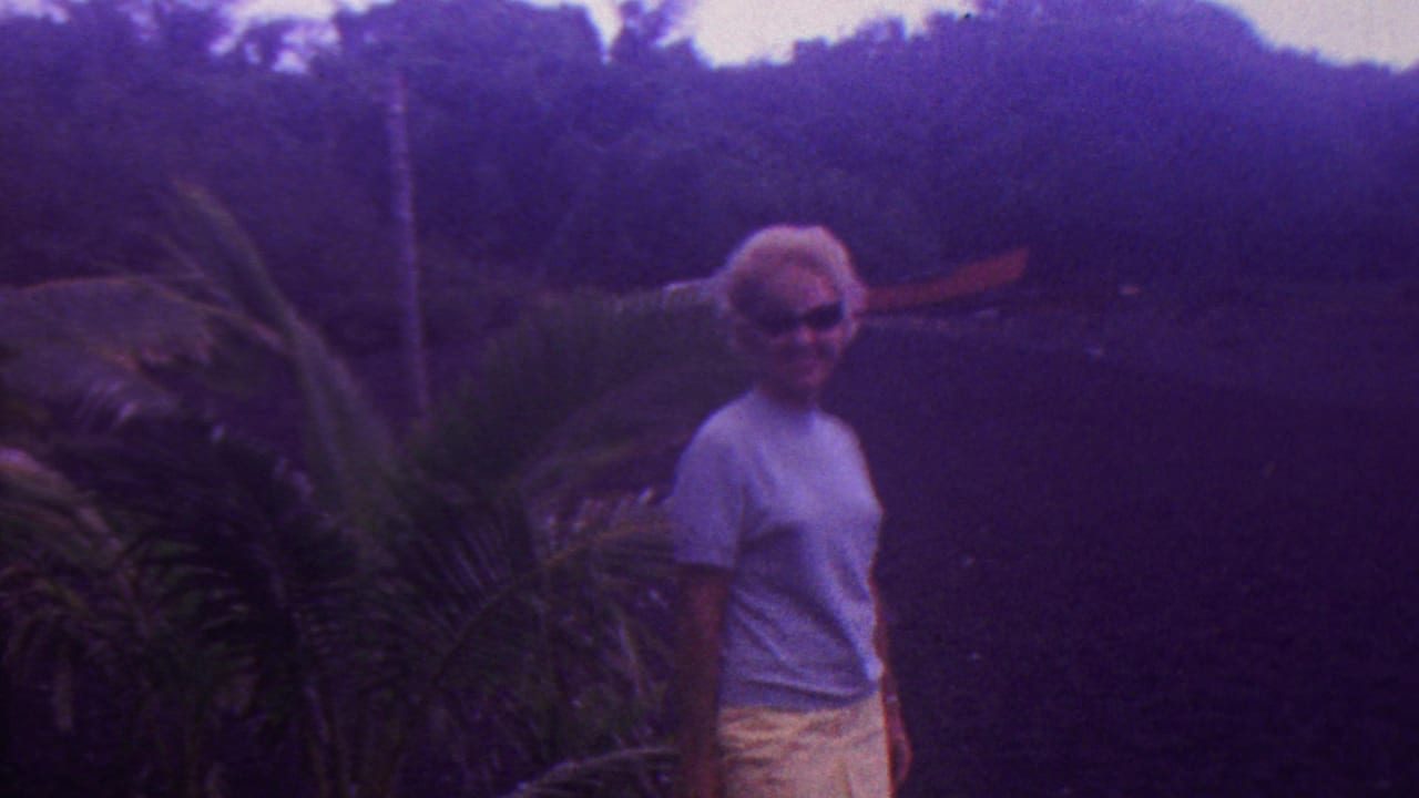 In supplied 1962 Hawaii home-movie footage, the woman remains posed outdoors beside tropical plants with dark foliage behind her.