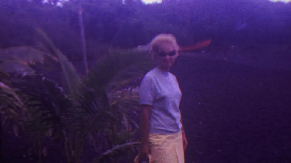 In the Hawaii home-movie sequence, the woman smiles toward the camera while standing near palm-like leaves and shaded vegetation.