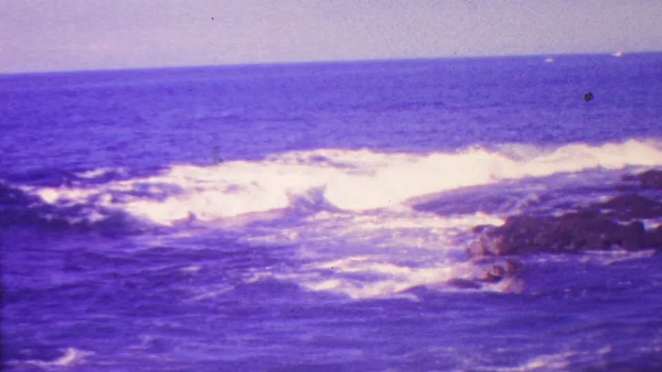 In supplied Hawaii footage, blue ocean water and white surf break against a dark rocky shoreline under aged purple-blue film color.