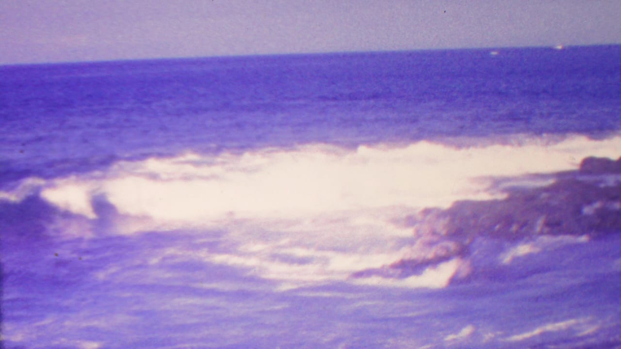 1962 Hawaii 8mm frame showing white waves breaking against dark coastal rocks under a blue-purple ocean horizon.