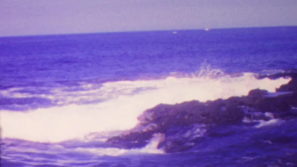 1962 Hawaii 8mm frame with a wave striking dark volcanic-looking rocks and spreading white foam across the shoreline.