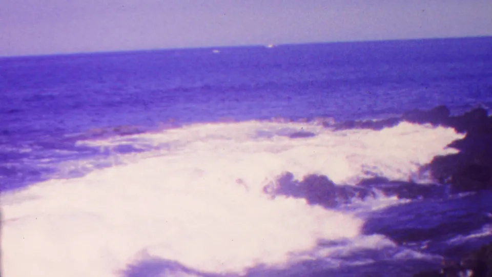 1962 Hawaii 8mm frame showing foamy surf washing around dark shoreline rocks with open water beyond.
