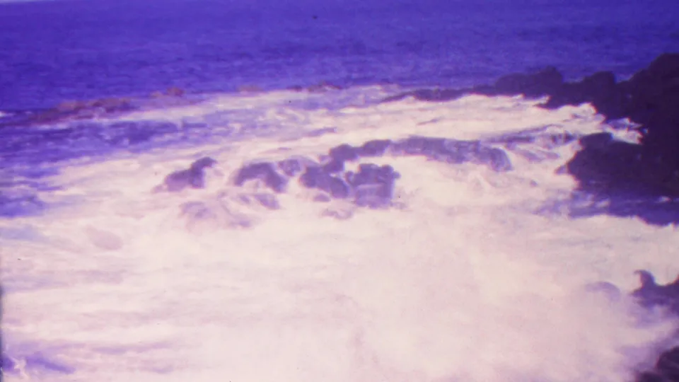 1962 Hawaii 8mm frame showing broad white surf rolling over dark coastal rocks beneath a purple-blue sea horizon.