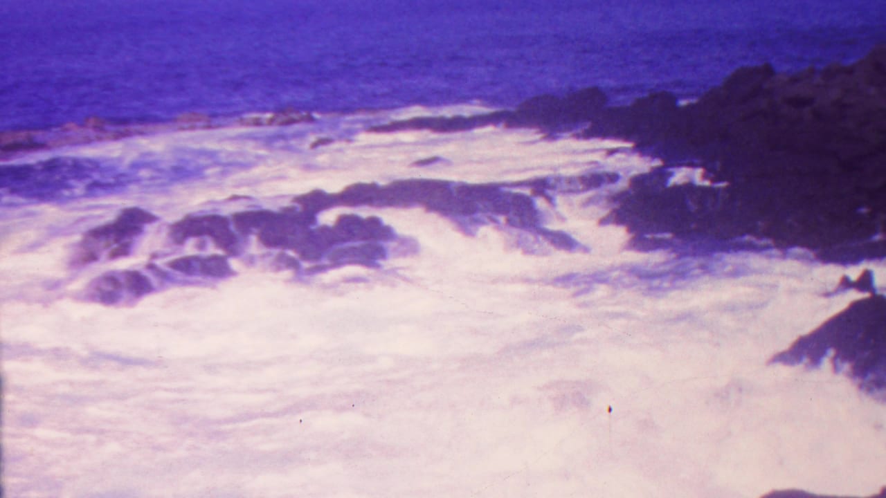 1962 Hawaii 8mm frame showing foamy waves rushing around dark coastal rocks with a strong magenta-purple film cast.