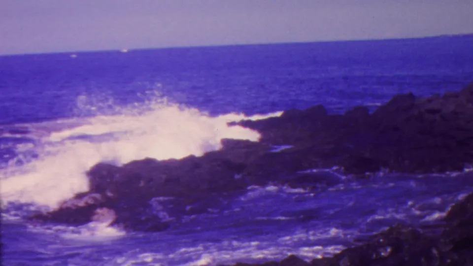 1962 Hawaii 8mm frame capturing a wave impact against dark shoreline rocks with spray rising above the blue water.