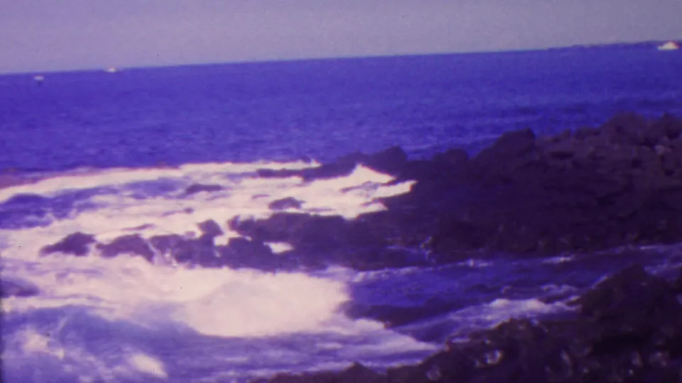1962 Hawaii coastal frame showing churned white surf below dark rocks, open ocean, and a level horizon under a bright sky.
