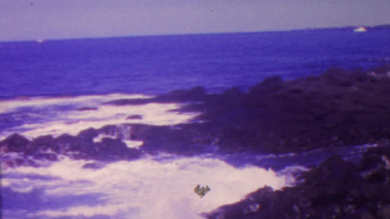1962 Hawaii 8mm home-movie frame showing white surf breaking around dark volcanic rocks beneath a blue ocean horizon and pale sky.