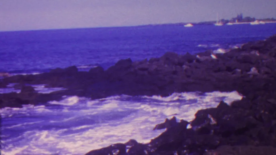 1962 Hawaii 8mm frame showing surf washing over dark volcanic rocks, open blue water, and faint distant boats near the horizon.