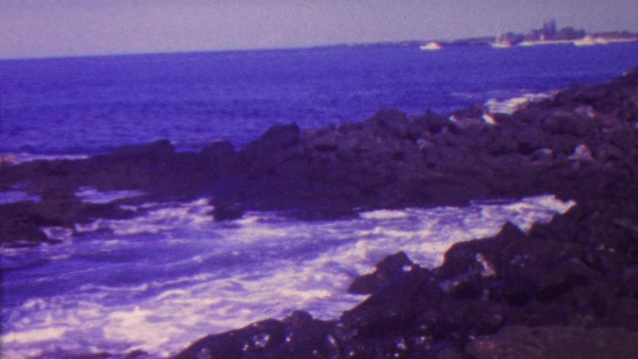 1962 Hawaii 8mm frame showing surf breaking across dark volcanic rocks, with a distant coastal strip and small boats visible offshore.