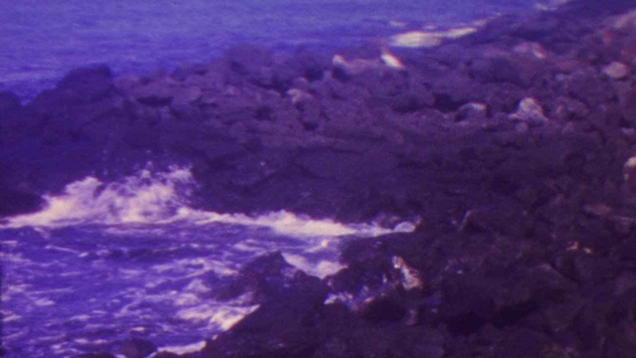 1962 Hawaii home-movie frame with white surf hitting dark shoreline rocks and open blue-purple ocean beyond.