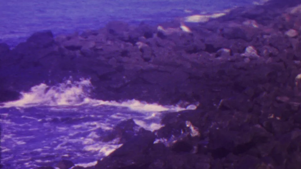 1962 Hawaii home-movie frame with white surf hitting dark shoreline rocks and open blue-purple ocean beyond.