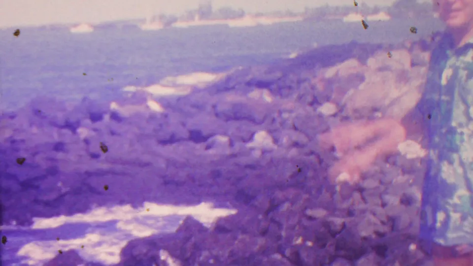 1962 Hawaii 8mm frame showing a partially visible man in a blue floral shirt gesturing toward waves breaking below the rocky shore.