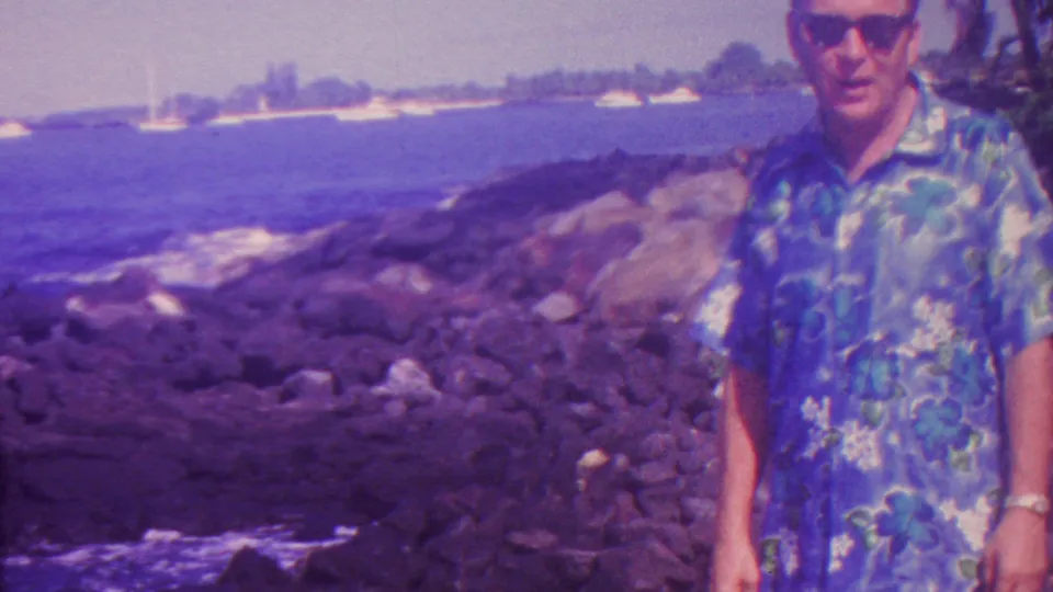 1962 Hawaii home-movie frame showing an adult man in sunglasses and a blue floral Hawaiian shirt standing beside dark rocks, surf, and distant sailboats.