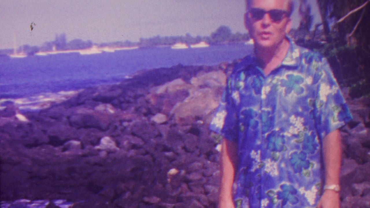 In 1962 Hawaii home-movie footage, an adult man in sunglasses and a blue floral shirt stands near dark coastal rocks with surf and a distant shoreline behind him.
