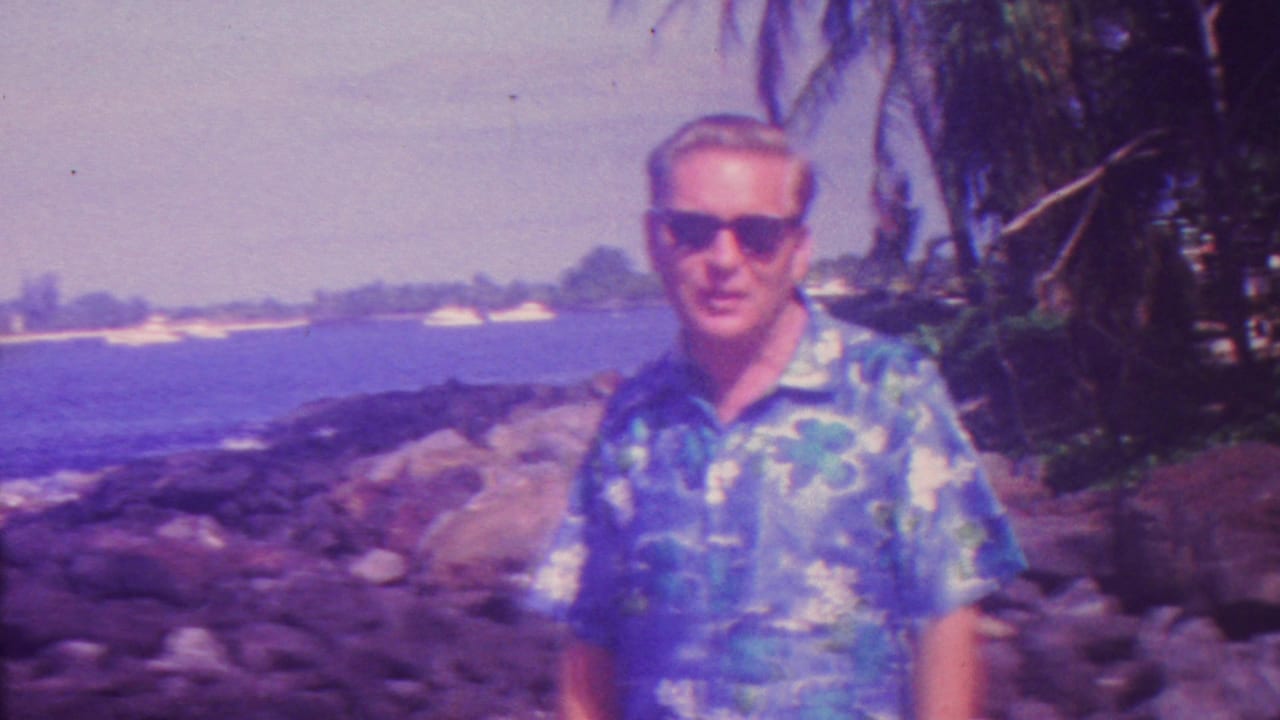 Man in sunglasses stands near rocks and palm foliage with blue ocean behind him.