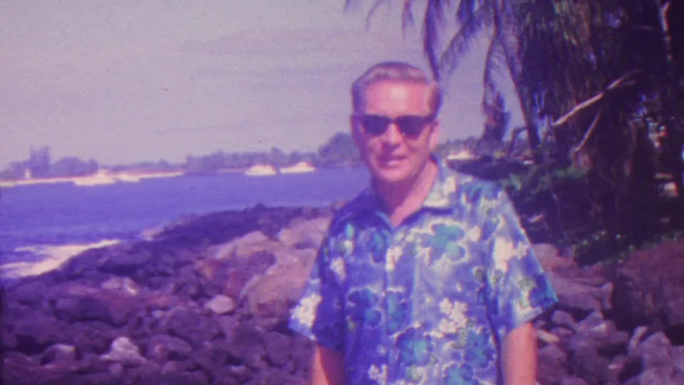 A closer 1962 Hawaii frame shows the man in a blue floral shirt and sunglasses beside dark lava-like rocks, breaking surf, and overhanging palm foliage.