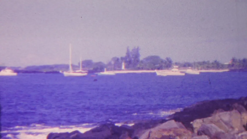 The final frame shifts away from the man to a Hawaii coastal view with sailboats offshore, low waves, foreground rocks, and a distant lighthouse-like vertical structure.