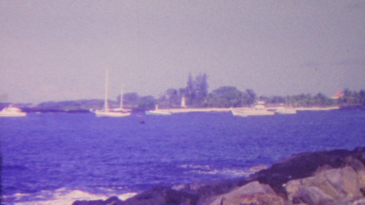 1962 Hawaii 8mm frame showing sailboats and small vessels on blue coastal water with rocks in the foreground and a low tropical shoreline in the distance.