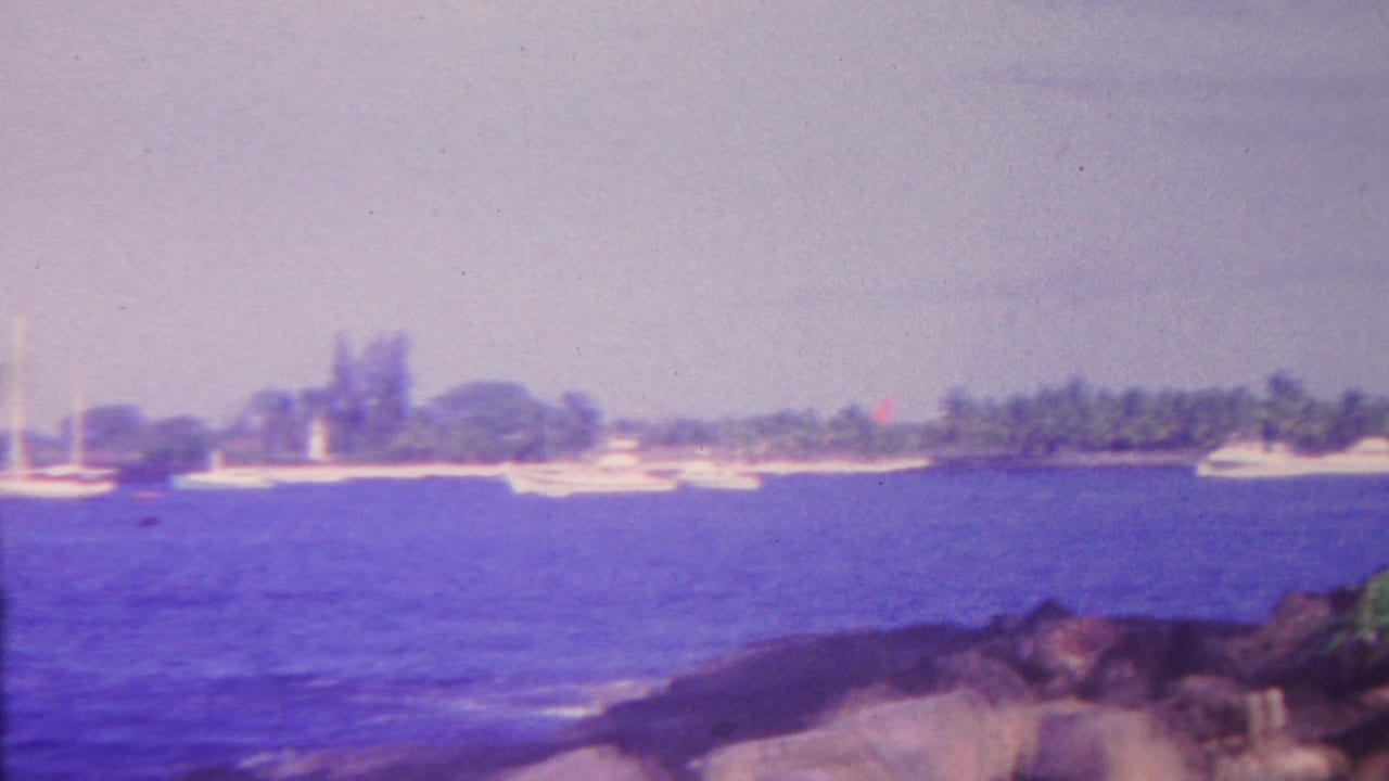 Boats sit offshore with rocks in the foreground and a low shoreline behind them.