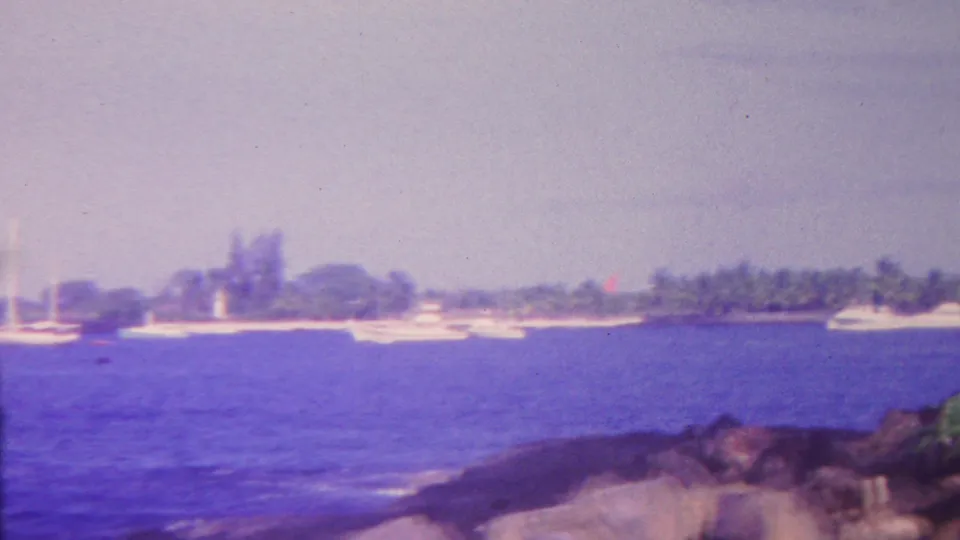 1962 Hawaii home-movie frame with anchored sailboats and small vessels near a distant tropical shore, seen over dark rocks at the water's edge.
