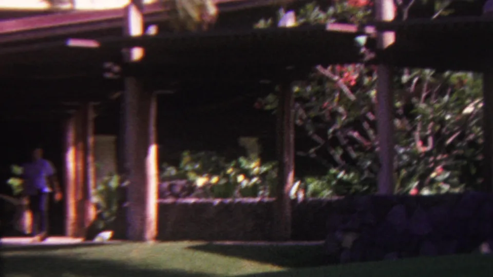 The camera looks across a covered patio area with wooden supports, stone walling, lawn, and dense tropical landscaping in the Hawaii resort grounds.