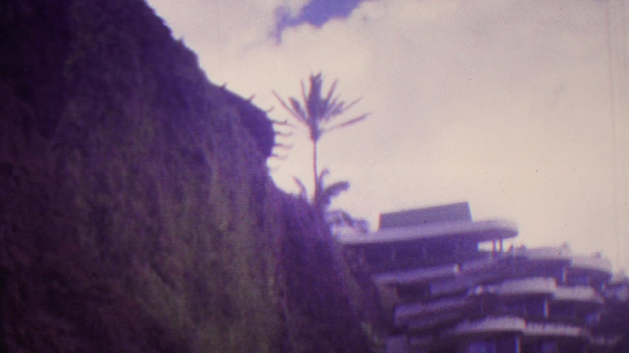 1962 Hawaii 8mm frame showing a steep vegetated hillside, a palm tree, and part of a curved multi-story building under pale clouds.