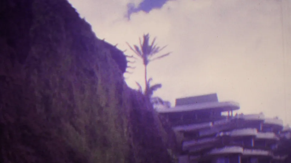 1962 Hawaii 8mm frame showing a steep vegetated hillside, a palm tree, and part of a curved multi-story building under pale clouds.
