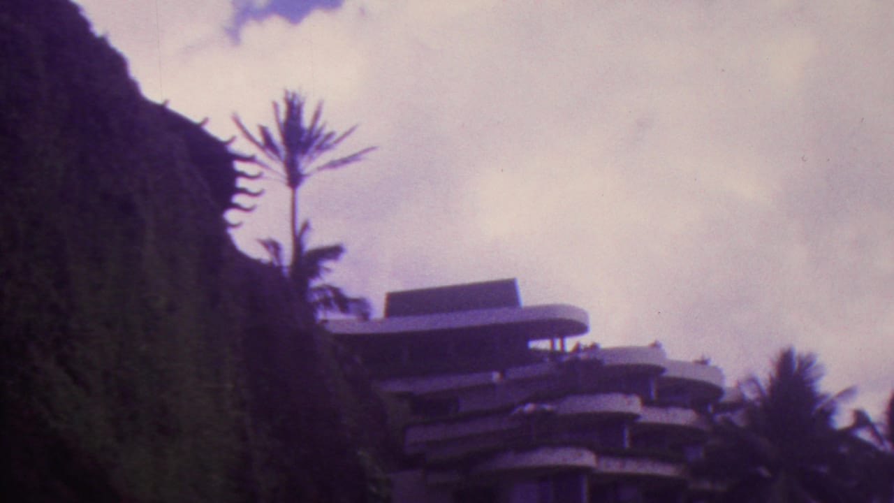 1962 Hawaii 8mm frame with a dark cliff edge, silhouetted palm, and modern tiered building visible against a cloudy sky.