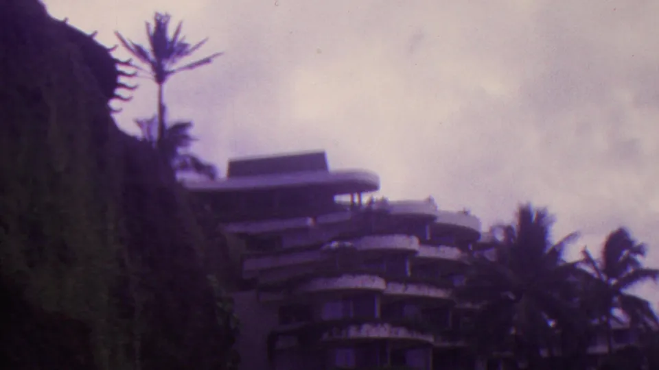 1962 Hawaii 8mm frame showing a curved multi-story building at the base of a steep green hillside with palm trees and hazy clouds behind it.