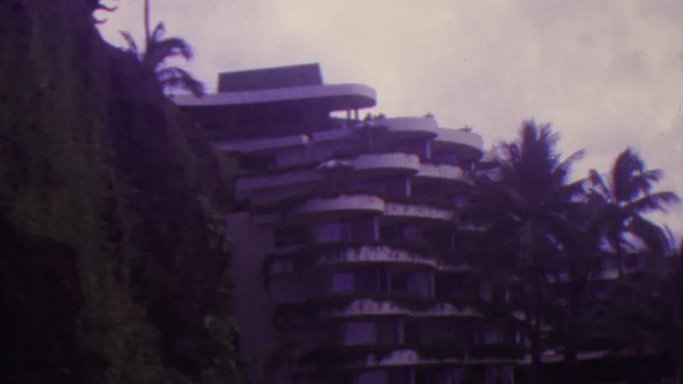 1962 Hawaii 8mm frame with a multi-story curved building framed by a steep vegetated cliff on the left and palms on the right.