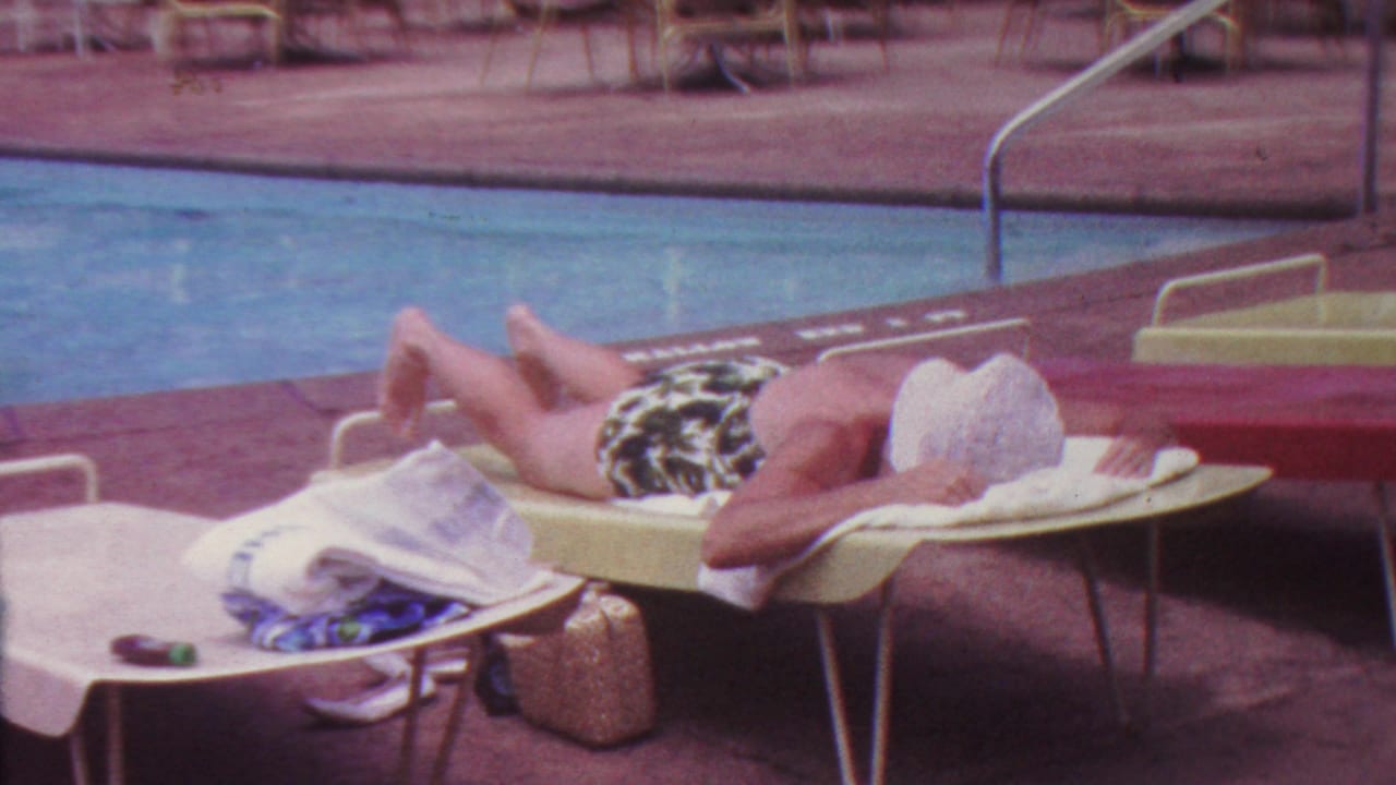 1962 Hawaii 8mm frame showing an adult woman in patterned swimwear reclining on a light lounge chair beside a swimming pool, with towels and deck furniture nearby.