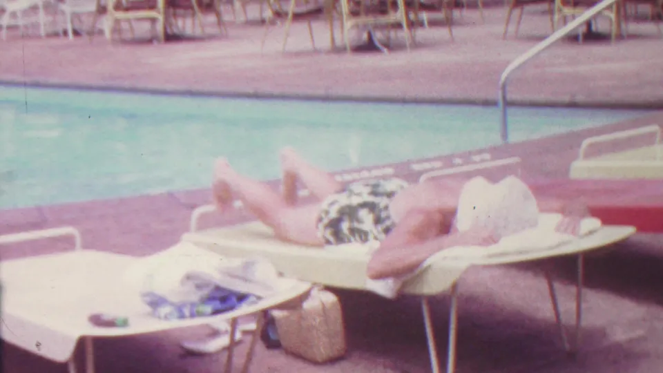 1962 Hawaii 8mm frame with a bright, overexposed view of an adult woman sunbathing on a lounge chair beside the pool, with towels and a small bag near the chair.