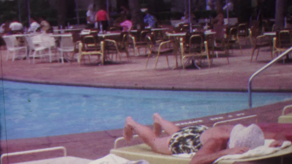 1962 Hawaii 8mm frame showing an adult woman reclining beside a swimming pool, with patio tables, chairs, and other poolside visitors in the background.